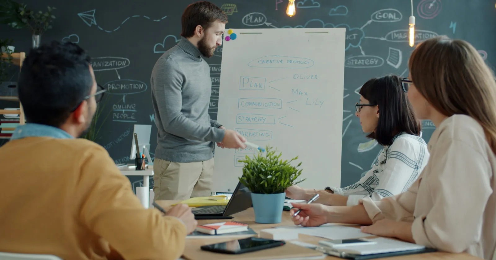 An engineer presenting a plan on a whiteboard to colleagues seated at a table with laptops